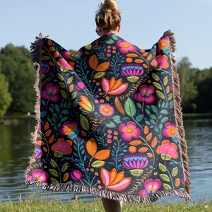 May include: A woven throw blanket with a colorful floral pattern on a black background. The blanket has fringe on all sides.