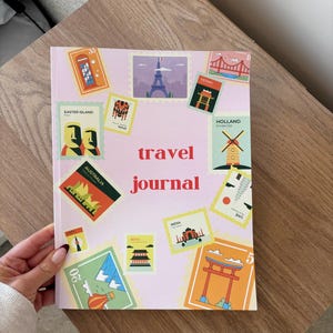 Puede incluir: Un diario de viaje rosa con un diseño colorido que presenta sellos con monumentos de todo el mundo. El diario tiene las palabras "travel journal" impresas en rojo en la portada.