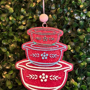 May include: A red hanging ornament shaped like a stack of three nesting bowls. Each bowl features a light blue floral pattern with leaves and a central flower. The ornament is suspended by a thin string attached to a small, round, light pink bead.