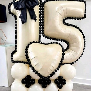 May include: A cream-coloured balloon display forming the number 15, with black beaded edging and accents. The number 1 is topped with a black bow. A heart-shaped balloon is also present. The base is made of white and black balloons. A celebratory decoration.