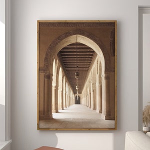 Op de afbeelding: Een ingelijste foto van een lange, smalle gang met gewelfde doorgangen en een betegelde vloer. De gang wordt verlicht door een enkele lamp die aan het plafond hangt. De foto is in een houten lijst.