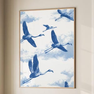 Puede incluir: Una impresión artística enmarcada que muestra flamencos azules volando contra un cielo nublado. La obra de arte se exhibe sobre un sillón beige con un cojín gris y una manta gris oscura. La habitación tiene una paleta de colores neutros.