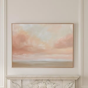 Puede incluir: Una pintura de una puesta de sol rosa y naranja sobre un cuerpo de agua tranquilo. La pintura está enmarcada en un marco de madera clara y cuelga sobre una chimenea de mármol blanco.