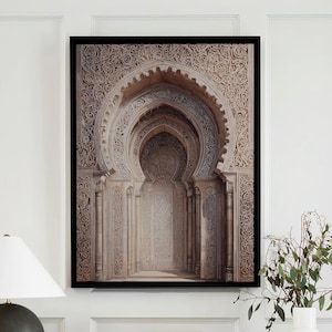 May include: A framed architectural print with an arched design hangs above a console table. A dark green lamp with a white shade, books, and a vase with greenery are on the table. The table has a marble top and black legs.