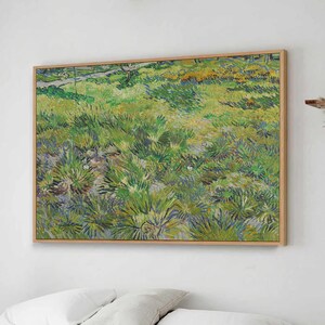 Op de afbeelding: Een ingelijst schilderij van een veld met groen gras, met een wit bed en een houten bank op de voorgrond. Het schilderij hangt aan een witte muur in een slaapkamer.