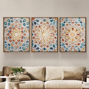 Op de afbeelding: Drie ingelijste aquarel kunstwerken met geometrische patronen in tinten oranje, teal en geel. Het kunstwerk wordt tentoongesteld boven een beige bank, waardoor een warme en uitnodigende woonkamerscène ontstaat.