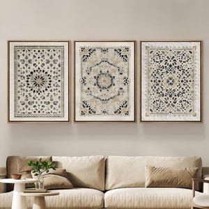 Op de afbeelding: Drie ingelijste wandkunstwerken met ingewikkelde, symmetrische patronen in beige, zwart en goudtinten. De kunstwerken worden tentoongesteld boven een beige bank met bijpassende kussens, wat zorgt voor een samenhangende en elegante woonkameresthetiek.