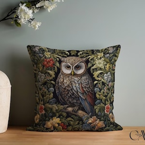 May include: Square decorative pillow featuring a detailed illustration of an owl perched on a branch, surrounded by floral elements. The pillow has a dark background with colorful flowers and foliage. A cream-colored vase with white flowers is to the left.