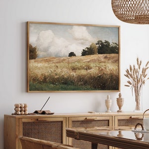 Puede incluir: Una pintura de paisaje enmarcada que representa un campo con árboles en el fondo. La pintura está colgada en una pared blanca sobre un armario de madera con puertas de ratán tejido. Hay flores secas y un jarrón sobre la mesa delante del armario.