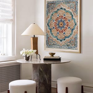 K&ouml;nnte beinhalten: Ein Raum mit einem runden Marmortisch, einem zylindrischen Sockel und zwei wei&szlig;en Ottomanen. An der Wand h&auml;ngt ein gerahmtes Kunstwerk mit einem detaillierten, farbenfrohen Design. Eine Holzlampe mit beigem Schirm und wei&szlig;e Blumen stehen auf dem Tisch.