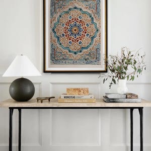 K&ouml;nnte beinhalten: Ein gerahmter Druck mit einem aufwendigen, symmetrischen Design in Blau-, Rot- und Beiget&ouml;nen h&auml;ngt &uuml;ber einer Konsole. Der Tisch hat eine Marmorplatte, schwarze Metallbeine und ist mit B&uuml;chern, einer Lampe und einem kleinen dekorativen Objekt ausgestattet.