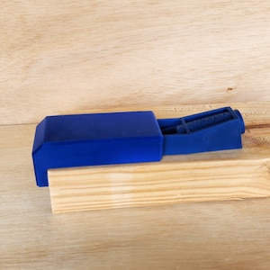 May include: A blue plastic jig for pocket holes, a woodworking tool for joining pieces of wood at an angle. The jig is resting on a wooden surface.