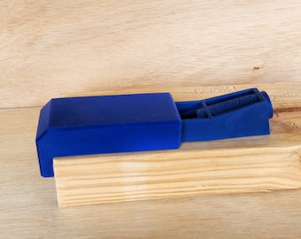 May include: A blue plastic jig for pocket holes, a woodworking tool for joining pieces of wood at an angle. The jig is resting on a wooden surface.