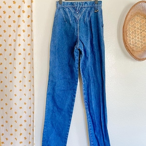 May include: Blue denim jeans with a high waist and a decorative back yoke. The jeans are hanging on a black hanger against a white wall. A straw hat and a floral curtain are in the background.