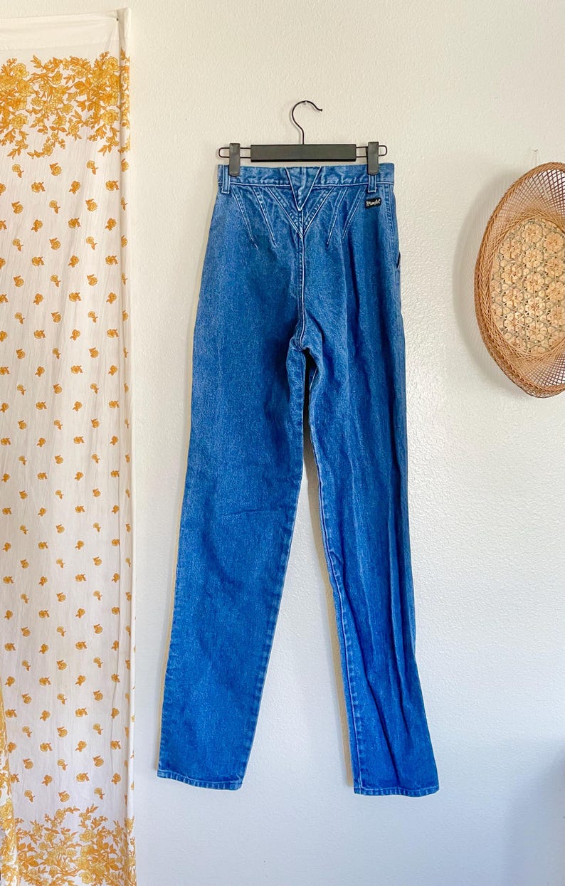May include: Blue denim jeans with a high waist and a decorative back yoke. The jeans are hanging on a black hanger against a white wall. A straw hat and a floral curtain are in the background.