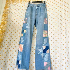 May include: Light blue denim jeans featuring wide side panels made of patchwork fabric. The panels include various patterned squares and white star shapes, along with embroidered lightning bolt designs running down the outer legs.