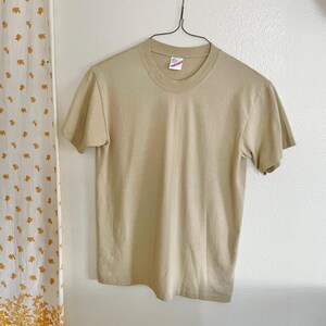 May include: A short-sleeved, tan-colored t-shirt hangs on a metal hanger against a white wall. The shirt has a crew neck and a tag is visible at the neckline. A patterned curtain is visible on the left side of the image.