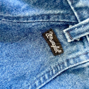 May include: Close-up of blue denim fabric with visible stitching and a black rectangular label. The label features the white text "Wrangler" in a cursive font, indicating the brand of the jeans.