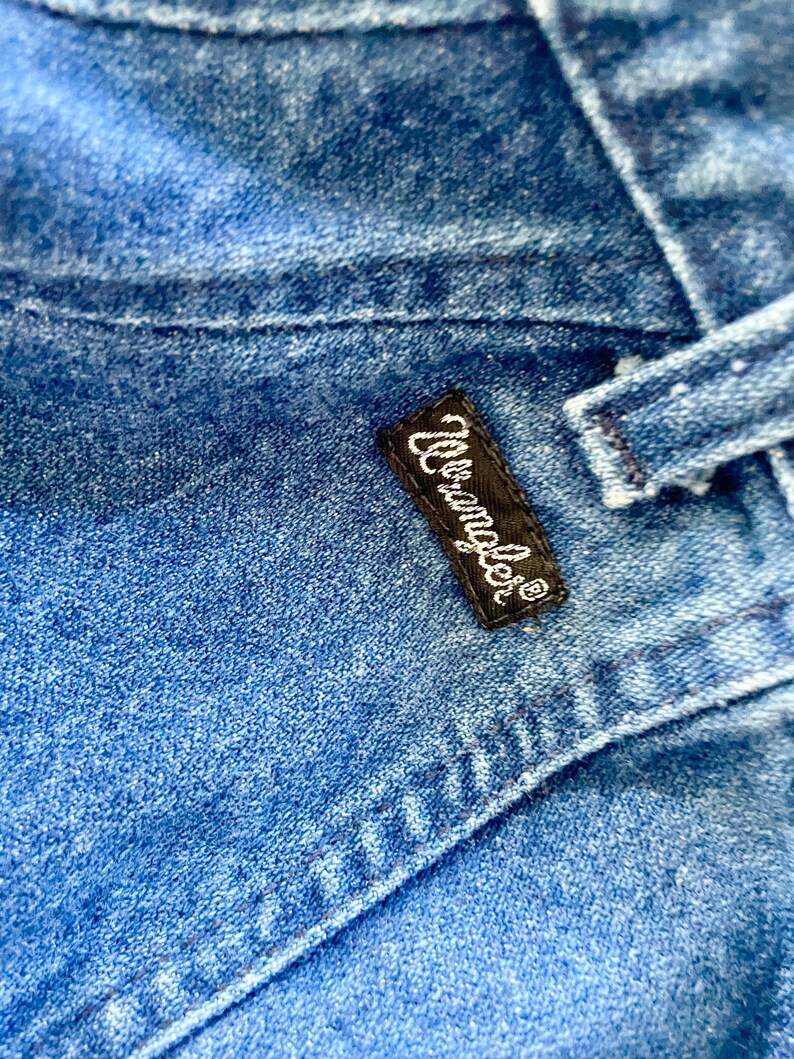 May include: Close-up of blue denim fabric with visible stitching and a black rectangular label. The label features the white text "Wrangler" in a cursive font, indicating the brand of the jeans.
