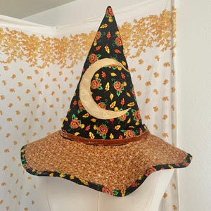May include: A pointed witch hat featuring a black fabric crown decorated with orange pumpkins and colorful autumn leaves, accented by a large gold crescent moon. The wide, wavy brim is covered in a brown floral print fabric with visible stitching.