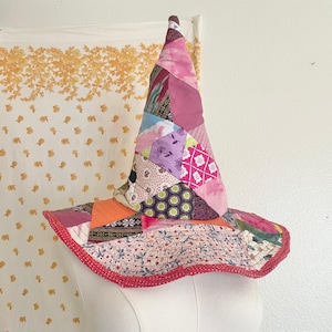 May include: A pointed hat constructed from a patchwork of various fabric scraps in shades of pink, purple, orange, and white. The hat features a wide, floppy brim with a red dotted trim and a conical crown made of triangular fabric pieces.