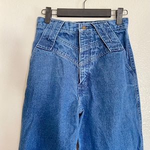 May include: Blue denim jeans with a high waist and distinctive yoke detailing. The jeans are suspended on a black hanger. The jeans feature a button closure and a classic five-pocket design. The background is neutral.