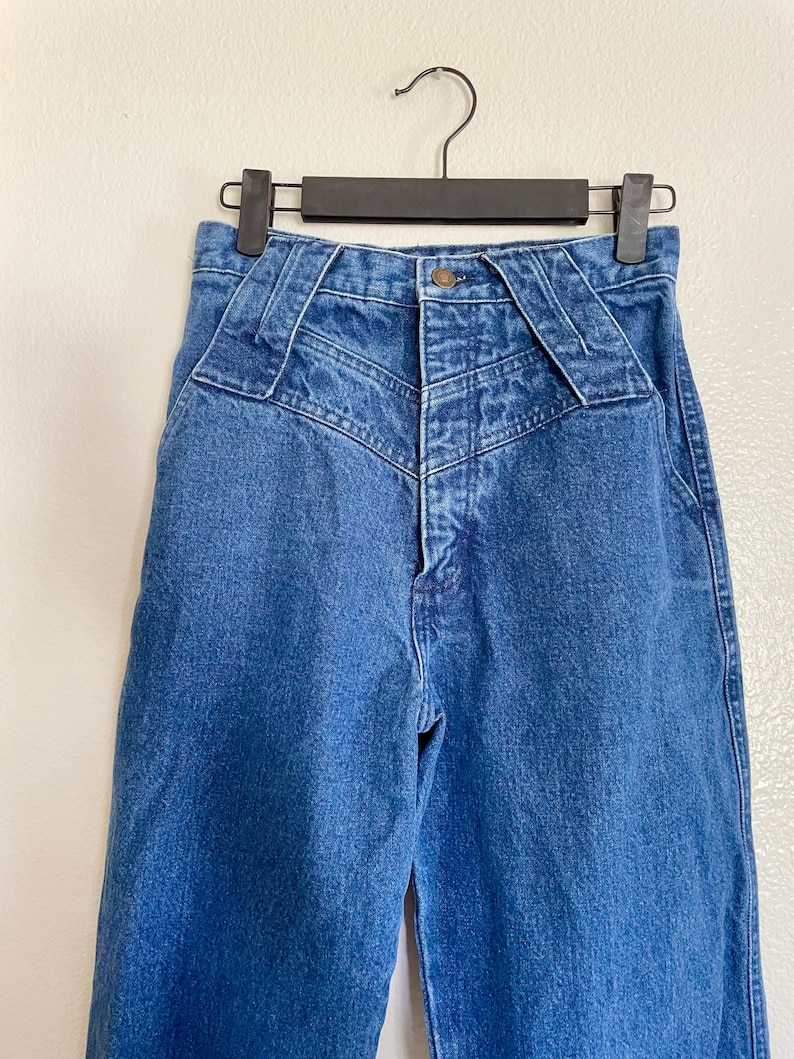 May include: Blue denim jeans with a high waist and distinctive yoke detailing. The jeans are suspended on a black hanger. The jeans feature a button closure and a classic five-pocket design. The background is neutral.