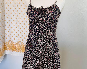 May include: A black floral print dress with spaghetti straps. The dress features a fitted bodice with a tie detail at the bust and a flared skirt. The fabric is covered in small pink and white flowers.