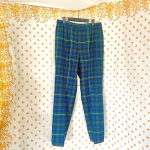 May include: Blue, green, and yellow plaid pants hanging on a black hanger. The pants have a straight leg cut and a button closure. The background is a white curtain with a gold floral pattern.