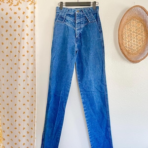 May include: Blue denim jeans with a high waist and a distinctive yoke design. The jeans are suspended on a black hanger. The background includes a woven wall decoration. The jeans are approximately 100 cm in length.
