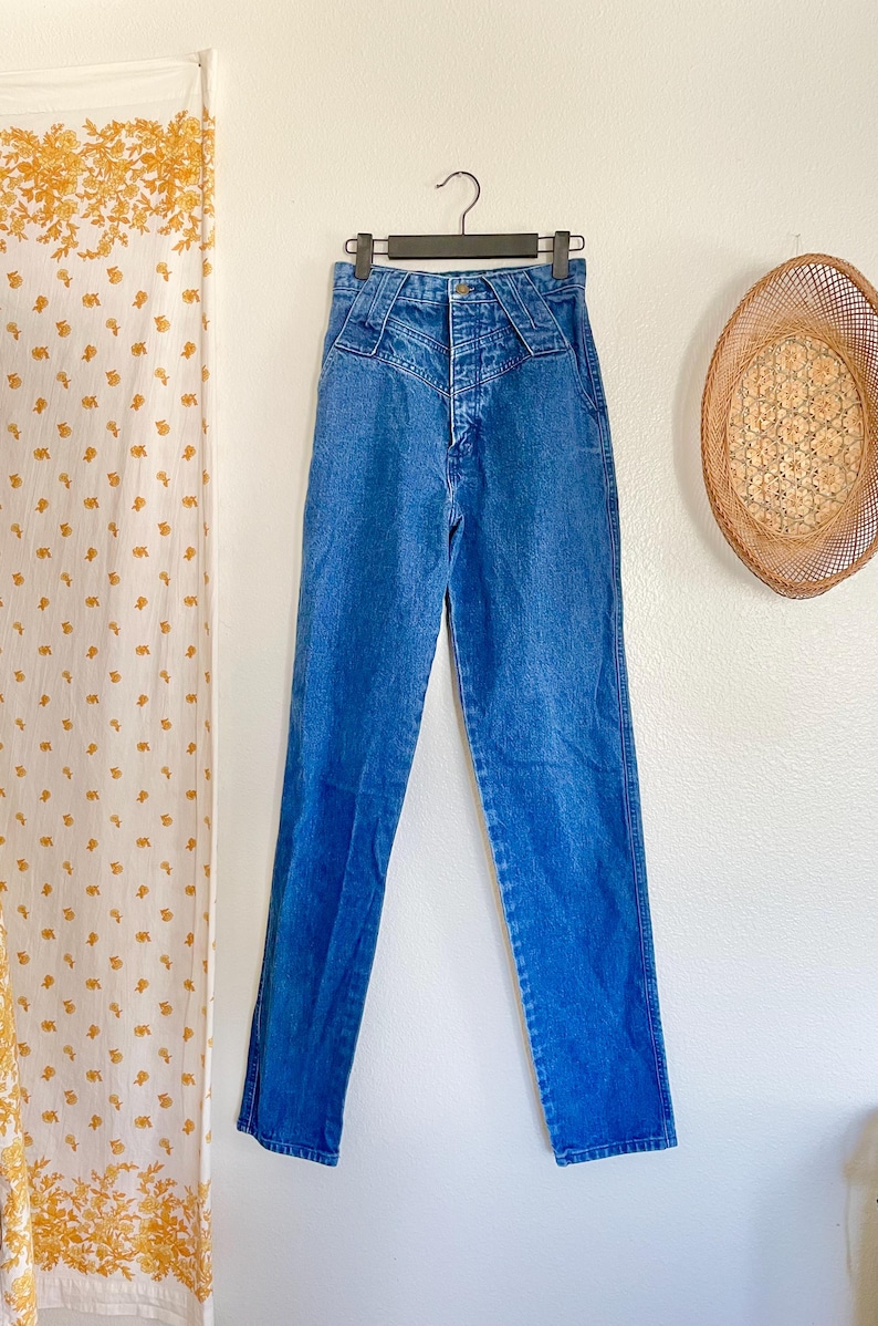 May include: Blue denim jeans with a high waist and a distinctive yoke design. The jeans are suspended on a black hanger. The background includes a woven wall decoration. The jeans are approximately 100 cm in length.