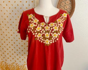 May include: A red short-sleeved blouse with a round neckline and intricate floral embroidery in white, yellow, and green. The blouse is displayed on a mannequin, showcasing its design and fit. The embroidery is concentrated around the neckline.
