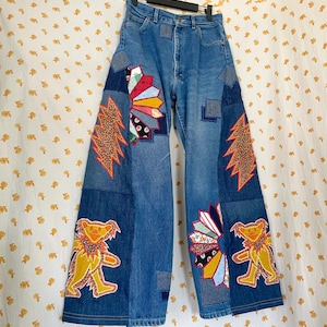 May include: High-waisted blue denim jeans featuring a wide-leg silhouette with various fabric patches. The jeans are decorated with colorful appliqués including two yellow dancing bears, two orange lightning bolts, and two multi-colored fans.