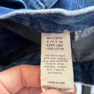 May include: Close-up of blue denim jeans with a white tag. The tag reads "NA729PW 3/4 X 36 A338-002 100% COTTON" and washing instructions. The jeans are made in the USA.