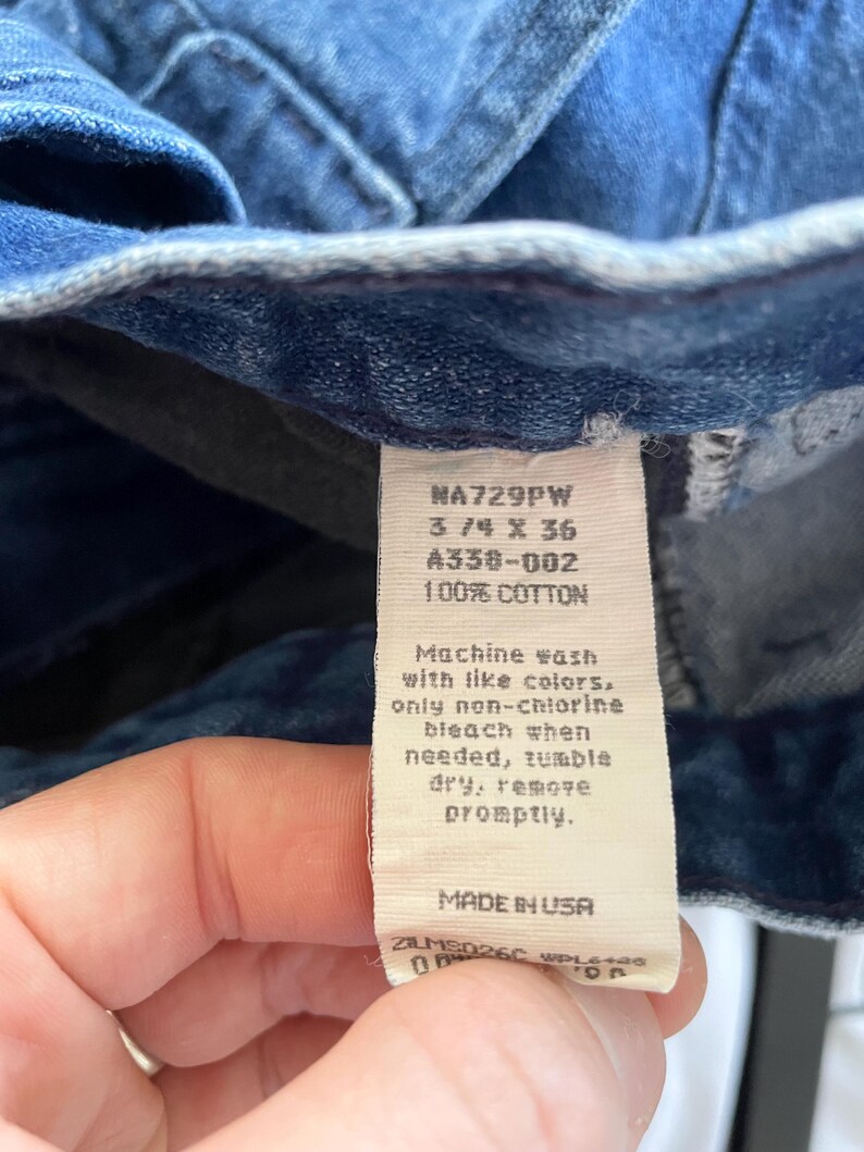 May include: Close-up of blue denim jeans with a white tag. The tag reads "NA729PW 3/4 X 36 A338-002 100% COTTON" and washing instructions. The jeans are made in the USA.