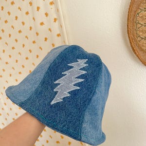 May include: A blue denim bucket hat with a white lightning bolt design. The hat has a slightly curved brim and a stitched construction. The lightning bolt is centered on the front of the hat.
