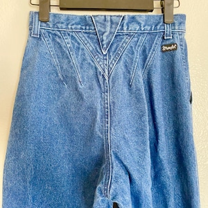 May include: Blue denim Wrangler jeans with a distinctive back yoke design. The jeans are suspended from a black hanger. The Wrangler logo is visible. These jeans have a classic, vintage style.