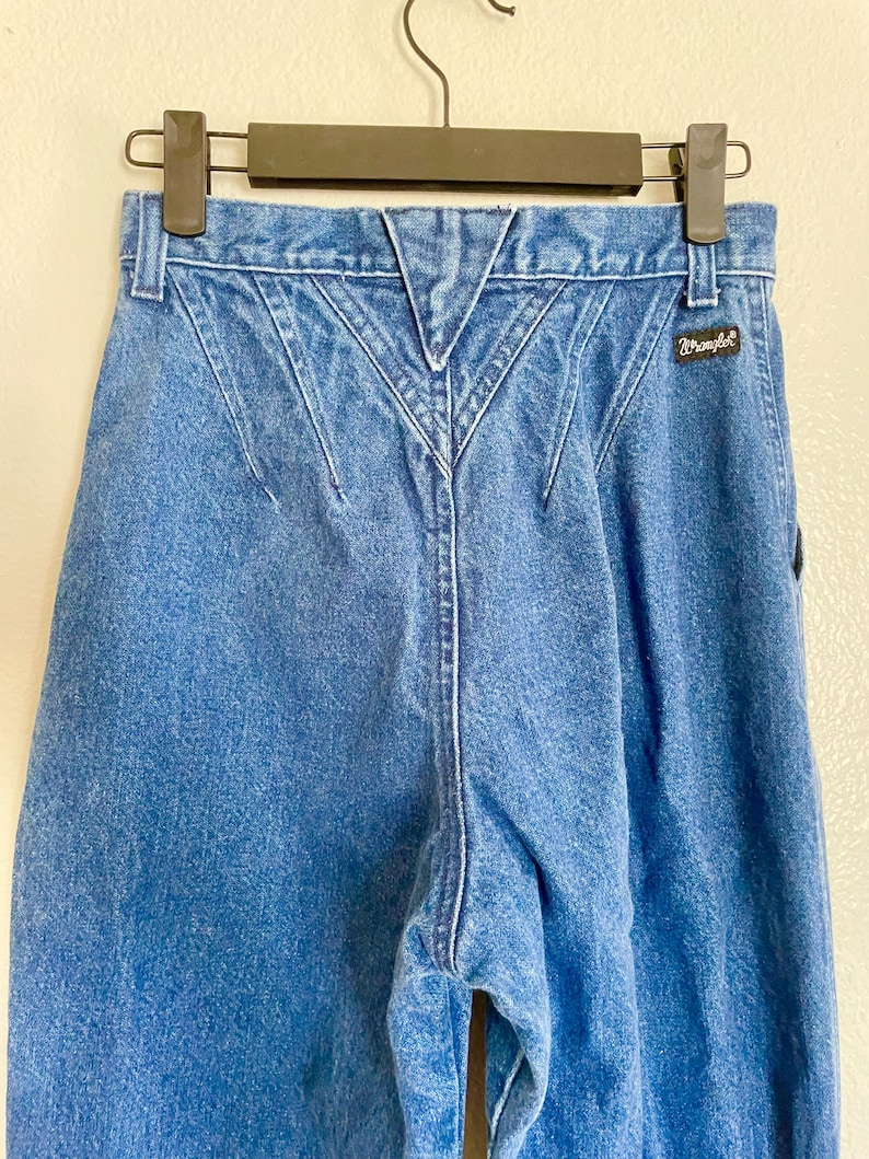 May include: Blue denim Wrangler jeans with a distinctive back yoke design. The jeans are suspended from a black hanger. The Wrangler logo is visible. These jeans have a classic, vintage style.