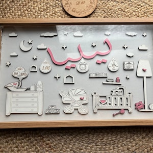 Peut inclure: Une œuvre d'art encadrée pour la chambre d'enfant avec un cadre en bois clair. L'œuvre présente un prénom de bébé en script arabe rose, ainsi que des découpes en bois de nuages, d'étoiles et d'articles pour bébés.