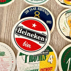 Pu&ograve; includere: Una collezione di sottobicchieri circolari in cartone con vari marchi Heineken, inclusi loghi rossi, bianchi e neri, un'illustrazione di un bicchiere di birra, grafiche di luppolo verde e testo in olandese come 200 Jaar Rijdende e Hopbel.