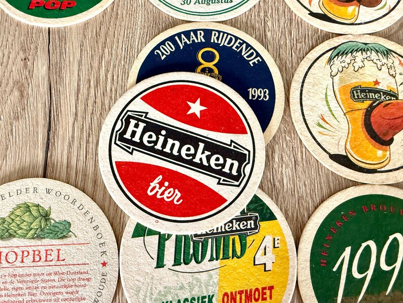 Pu&ograve; includere: Una collezione di sottobicchieri circolari in cartone con vari marchi Heineken, inclusi loghi rossi, bianchi e neri, un'illustrazione di un bicchiere di birra, grafiche di luppolo verde e testo in olandese come 200 Jaar Rijdende e Hopbel.