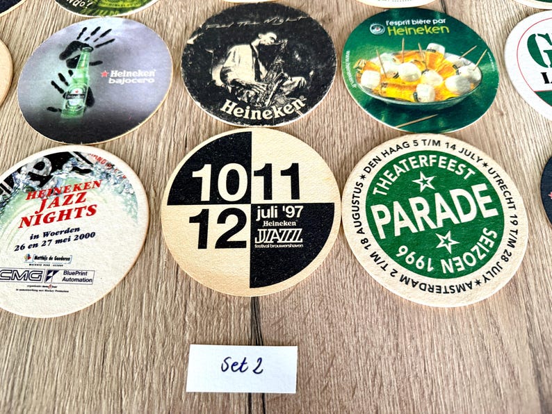 Pu&ograve; includere: Una collezione di sottobicchieri circolari in cartone con vari design grafici, tra cui il marchio Heineken, pubblicit&agrave; di festival jazz con date come 1997 e 2000 e un logo Theaterfeest Parade. Una piccola etichetta bianca riporta la scritta Set 2.