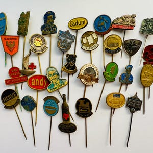 Könnte beinhalten: Eine Sammlung von Vintage-Anstecknadeln mit verschiedenen Designs und Farben. Die Anstecknadeln zeigen Bilder, Texte und Logos, darunter einen Bären, eine Flasche und den Text "Cadum". Die Anstecknadeln sind auf einer weißen Oberfläche angeordnet.