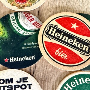 Pu&ograve; includere: Una collezione di sottobicchieri circolari in cartone con vari marchi Heineken. Un sottobicchiere mostra un cerchio rosso con il testo bianco Heineken e bier, mentre un altro mostra un grafico di una bottiglia verde con il testo Heineken e W każdym.