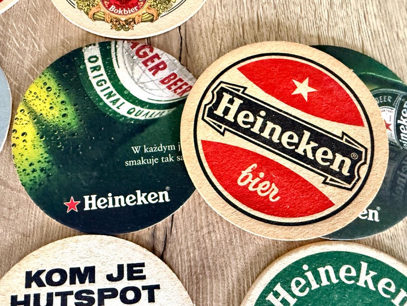 Pu&ograve; includere: Una collezione di sottobicchieri circolari in cartone con vari marchi Heineken. Un sottobicchiere mostra un cerchio rosso con il testo bianco Heineken e bier, mentre un altro mostra un grafico di una bottiglia verde con il testo Heineken e W każdym.