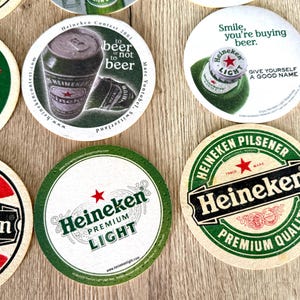 Pu&ograve; includere: Una collezione di sottobicchieri rotondi in cartone con vari marchi Heineken, inclusi loghi, lattine di birra e tappi a corona. I design includono testi come 'to beer or not to beer', 'Smile, you're buying beer' e 'Heineken Premium Light'.