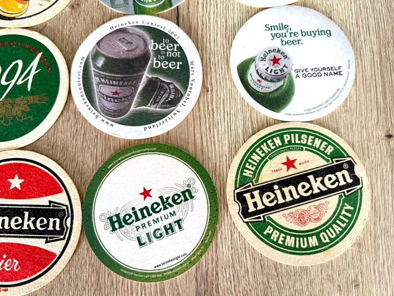 Pu&ograve; includere: Una collezione di sottobicchieri rotondi in cartone con vari marchi Heineken, inclusi loghi, lattine di birra e tappi a corona. I design includono testi come 'to beer or not to beer', 'Smile, you're buying beer' e 'Heineken Premium Light'.