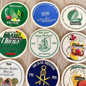 Pu&ograve; includere: Una collezione di sottobicchieri rotondi in cartone con vari disegni grafici, tra cui il marchio Heineken, temi musicali, testi commemorativi storici e illustrazioni di bicchieri di birra con accessori festivi come cappelli e occhiali.