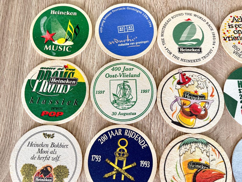 Pu&ograve; includere: Una collezione di sottobicchieri rotondi in cartone con vari disegni grafici, tra cui il marchio Heineken, temi musicali, testi commemorativi storici e illustrazioni di bicchieri di birra con accessori festivi come cappelli e occhiali.