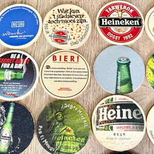 Pu&ograve; includere: Una collezione di dodici sottobicchieri circolari in cartone con vari marchi Heineken, grafiche promozionali e testo tra cui 'Be a Secret Agent for a Day', 'Bier!' e 'Heineken Bokbier'. I sottobicchieri mostrano colori, loghi e immagini diversi.